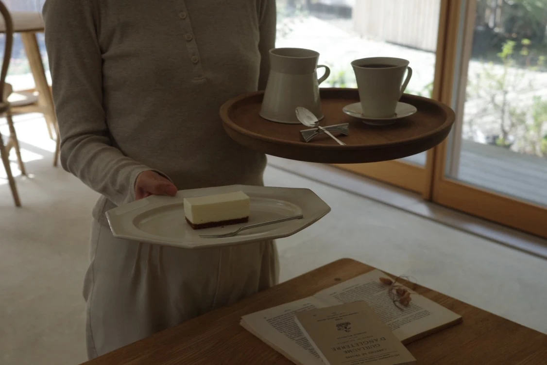 窓辺でレアチーズケーキと飲み物を運ぶ人の手元